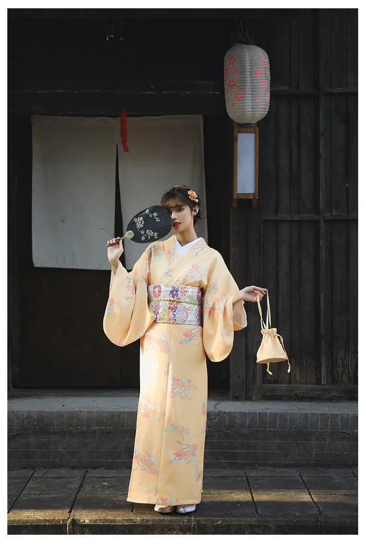 Woman in a retro orange Japanese kimono-style yukata with floral designs, holding a decorative fan and a small bag in a traditional setting.