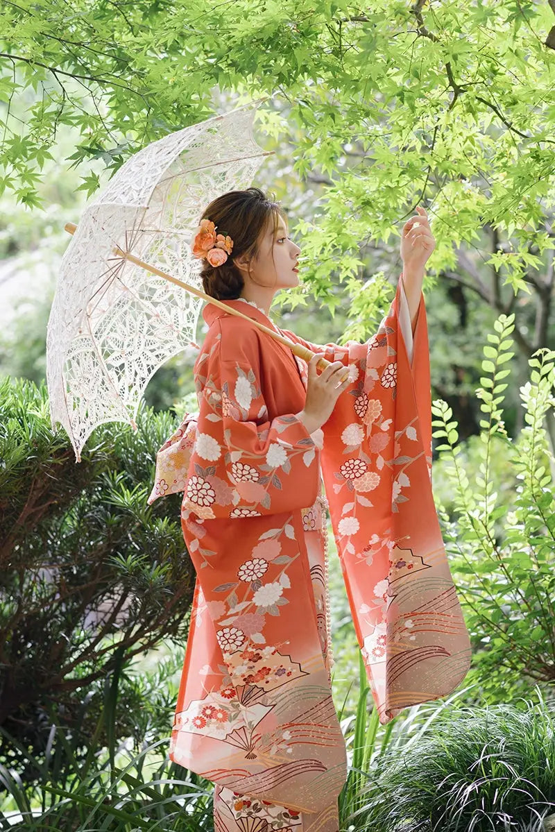 Side profile of a woman in an orange chrysanthemum-patterned kimono holding a lace parasol, set in a lush green outdoor scene.