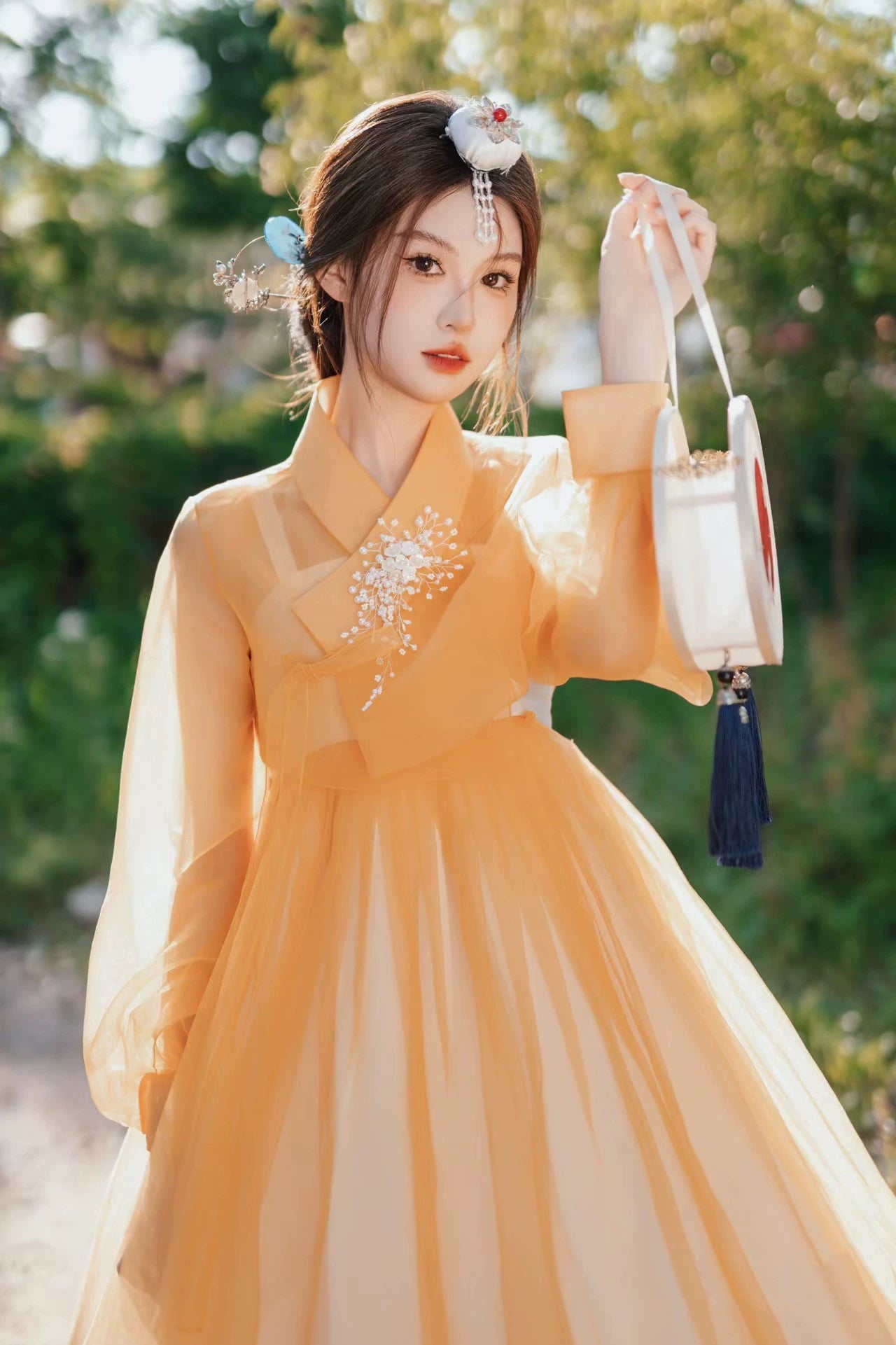 Graceful woman in an orange Yanbian Yanji Korean Hanbok dress, holding a round handbag with tassels, standing in a lush green outdoor setting.