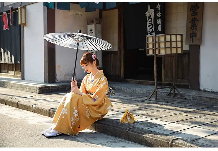 Woman in an orange floral kimono yukata sitting on a stone pavement with a traditional umbrella, surrounded by a serene Japanese street setting.