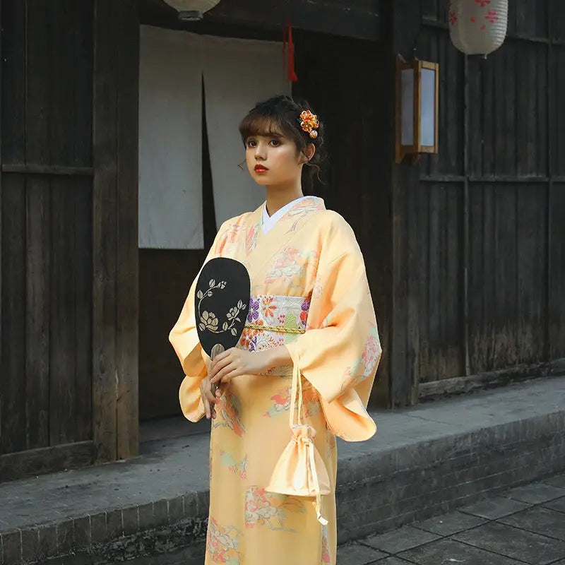 Close-up of a woman in a literary-style orange Japanese kimono-style yukata with floral patterns, holding a decorative fan.