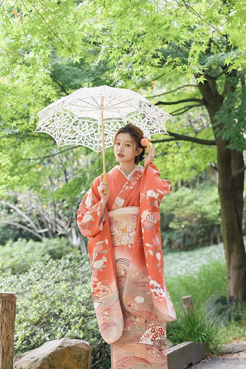 Full view of an orange chrysanthemum-patterned kimono with a lace parasol, styled for travel and photography in a lush garden.