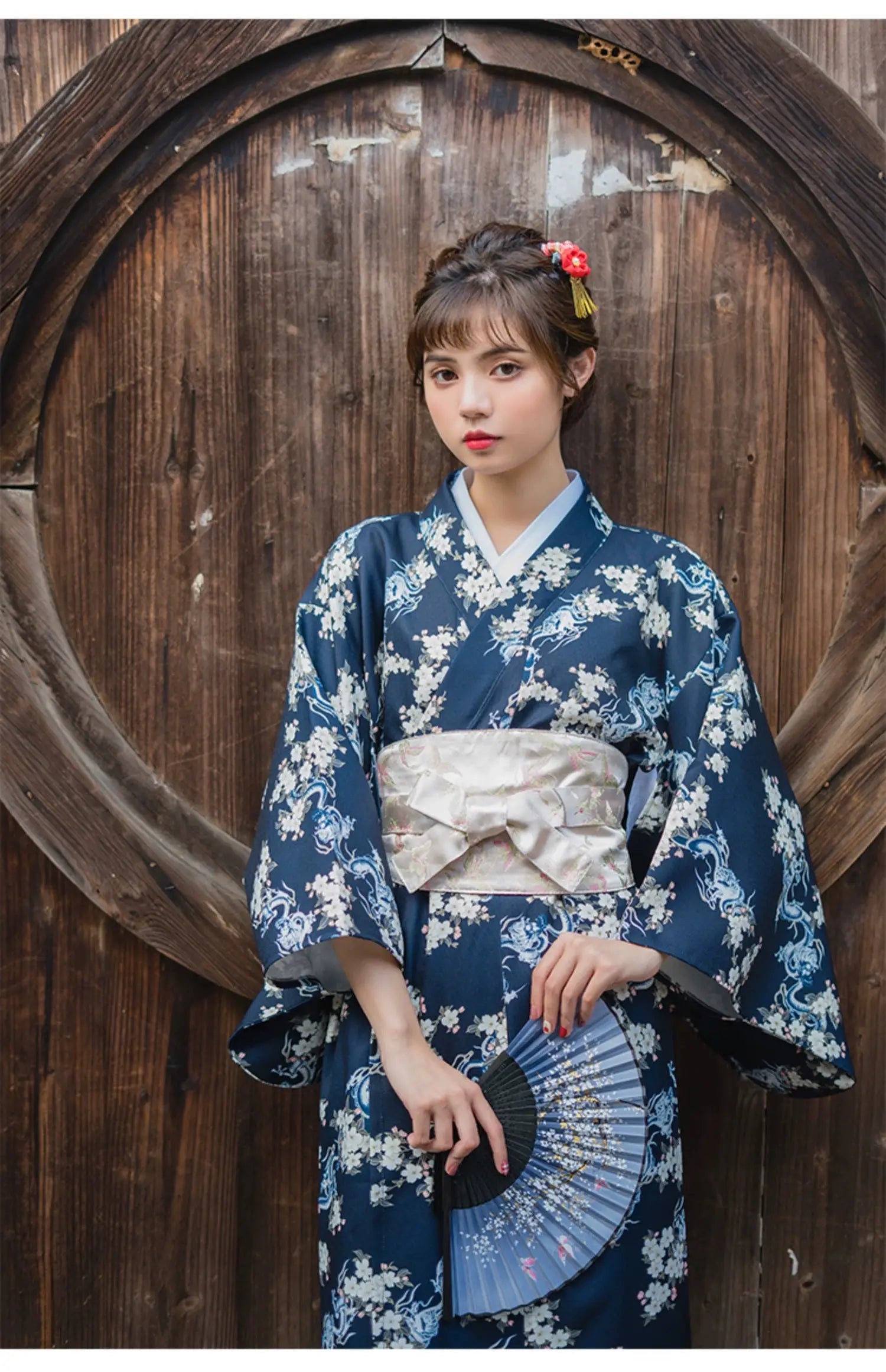 Woman in a blue floral yukata styled with a white brocade kimono belt seal, holding a fan and standing against a wooden background.