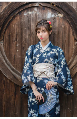 Woman in a blue floral yukata styled with a white brocade kimono belt seal, holding a fan and standing against a wooden background.