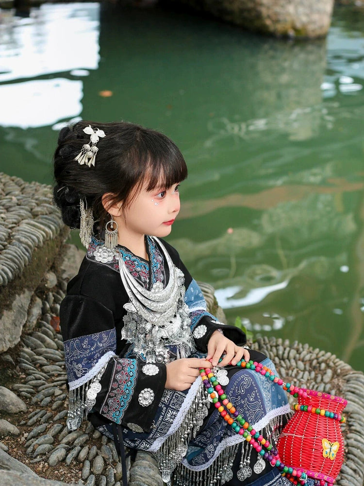 Girl in Miao blue Dai dance costume near water, holding colorful beads and red pouch, ideal for ethnic dance performances.
