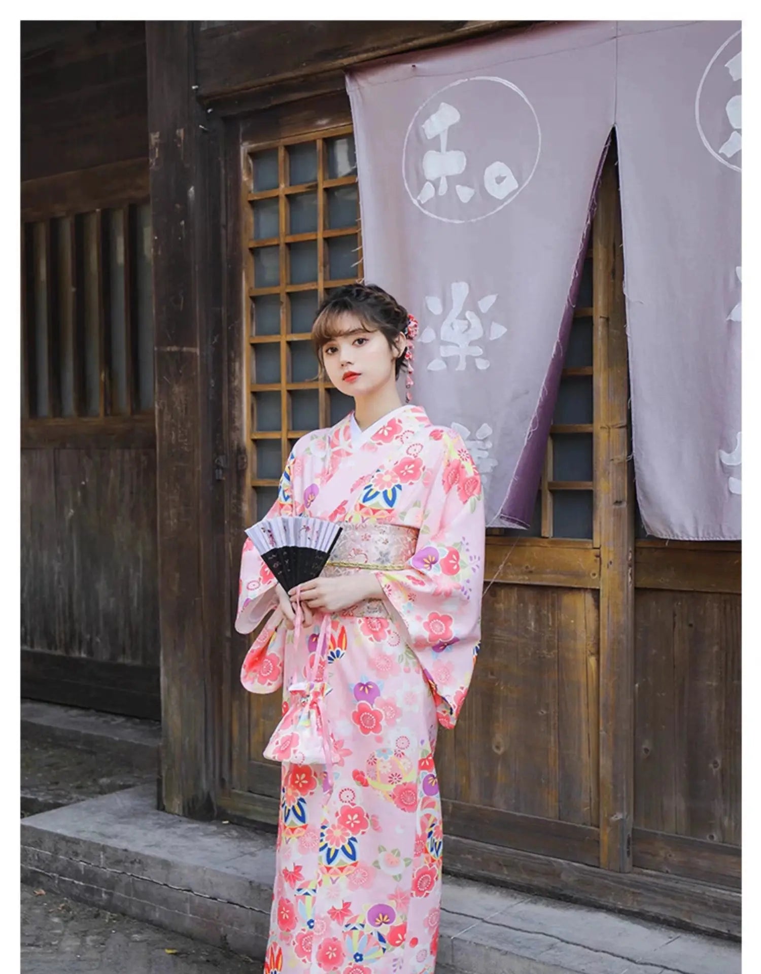 Woman in a pink floral kimono holding a fan, standing in front of a traditional Japanese wooden building with purple curtains.