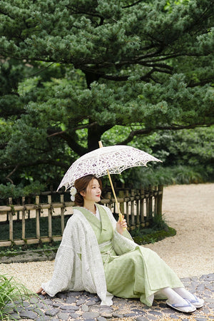 Model holding lace parasol in green kimono dress by a fence.