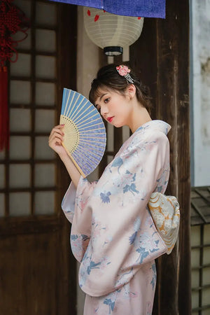 Side view of a woman in a pink yukata kimono with floral and crane designs, holding a fan, exuding a refined Japanese retro style.