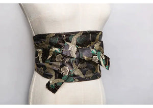 Black brocade kimono belt seal with gold and green crane and floral patterns, tied in a bow on a mannequin for a classic Japanese look.