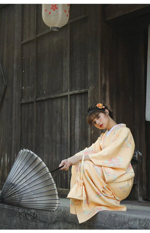 Woman seated in an orange Japanese kimono-style yukata with floral designs, holding a black umbrella, near a wooden wall.