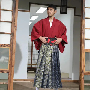 Man wearing a Japanese formal samurai costume with a red kimono and patterned brocade skirt, standing in a traditional wooden interior.