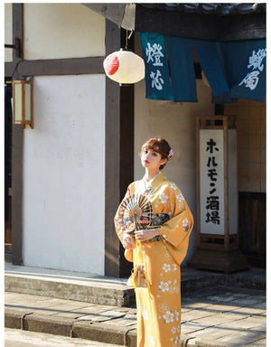 Woman in an orange floral kimono yukata holding a fan, gazing upward near a traditional Japanese building with lanterns and banners.