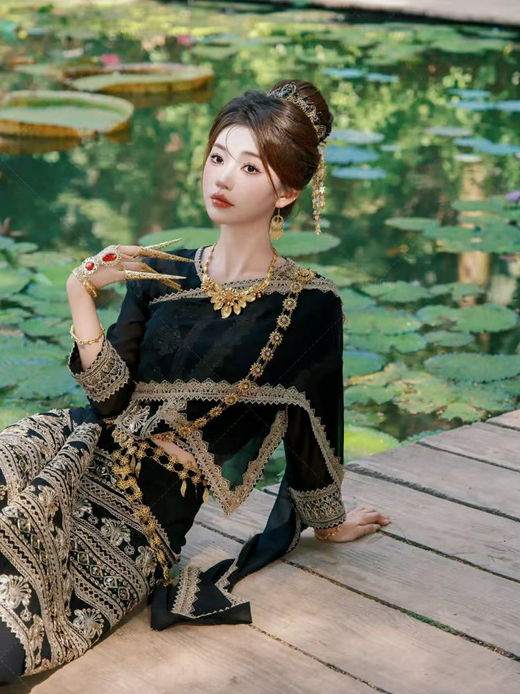 Model in Black&Gold Dai clothing female princess outfit reclining by lotus pond, showcasing full ethnic attire and accessories.