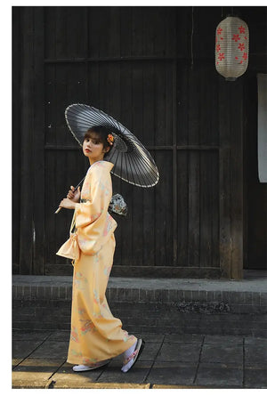 Woman in an orange Japanese kimono-style yukata with floral patterns, walking with a black umbrella in a traditional wooden street setting.