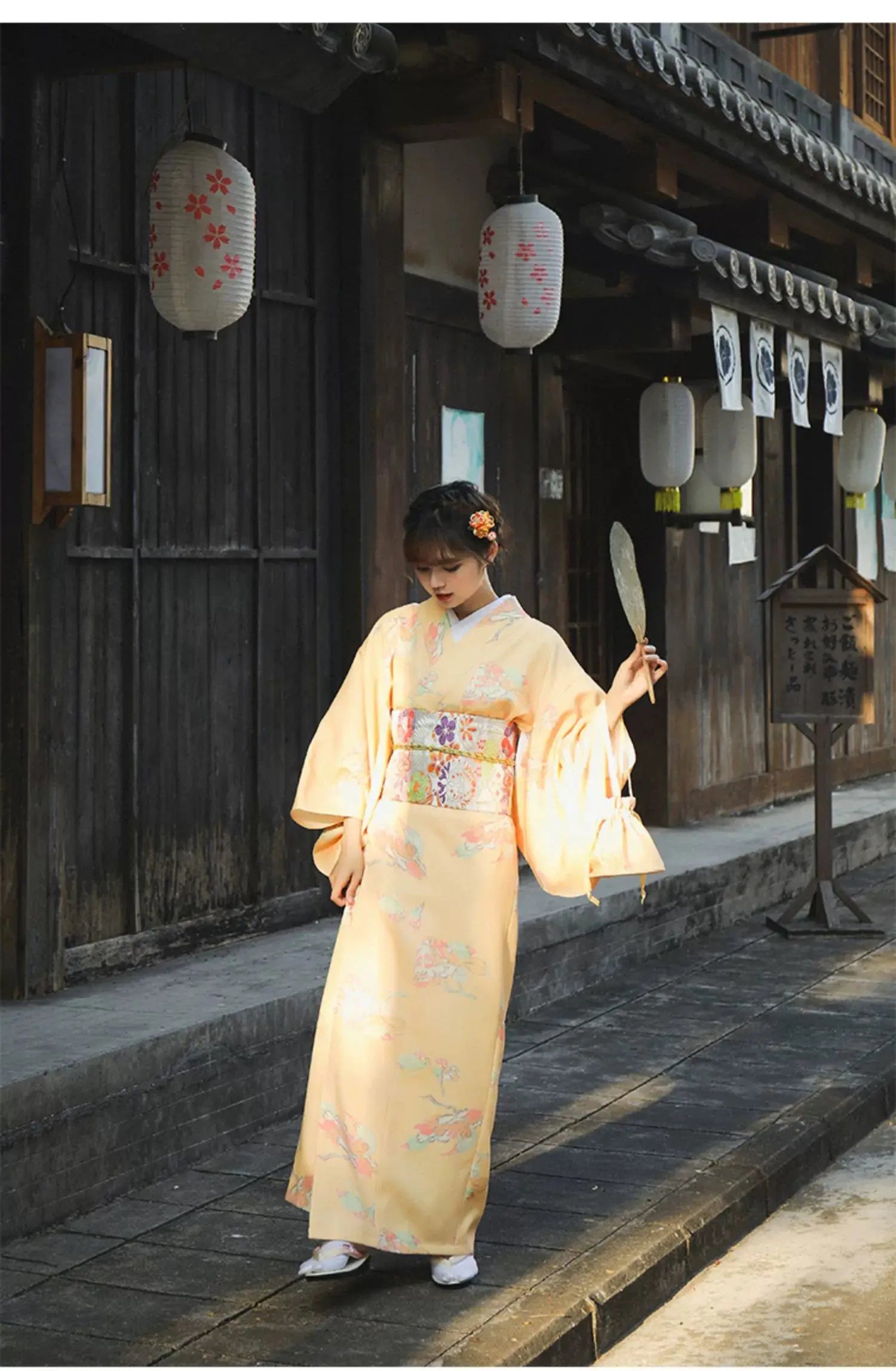 Woman in a formal orange Japanese kimono-style yukata with floral prints, holding a decorative fan in a traditional wooden street setting.