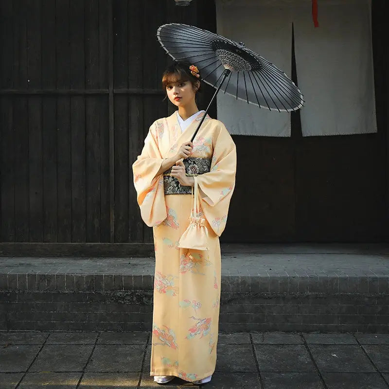 Woman dressed in a retro orange Japanese kimono-style yukata with floral prints, posing with a black umbrella in front of a wooden backdrop.