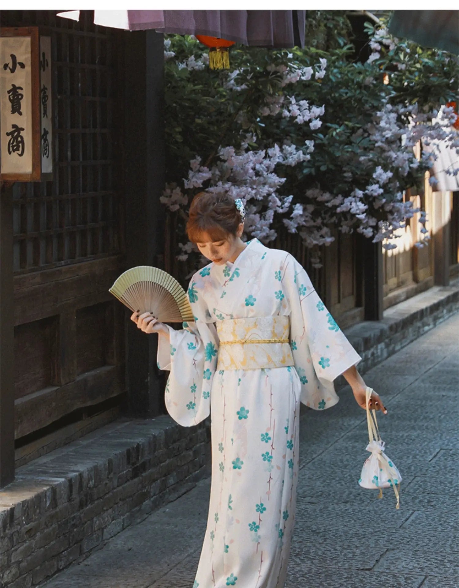 Beautiful girl wearing a white and blue floral Yukata Kimono Dress, holding a fan and a matching bag, set in a traditional Japanese street.