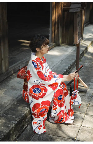 Woman in a white and red floral Japanese kimono dress, seated outdoors, holding a traditional accessory and showcasing the intricate design.