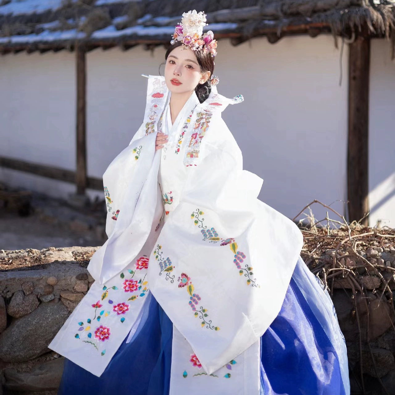 Bride in white Korean hanbok with embroidery, posing outdoors in sunlight.