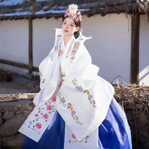 Bride in white Korean hanbok with embroidery, posing outdoors in sunlight.