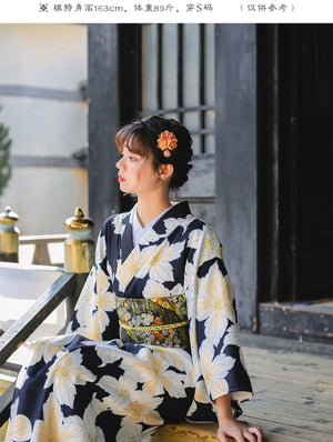 Woman wearing a Japanese kimono dress with a floral pattern, seated outdoors, styled with a traditional obi belt for a festive look.