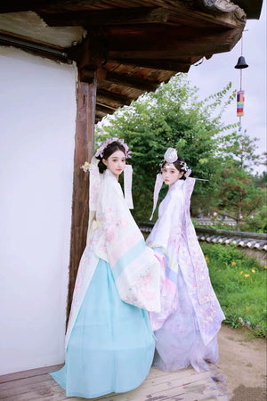 Two brides in Muzhi Korean embroidered wedding dresses, posing in a serene outdoor setting with delicate floral embroidery and pastel hues.