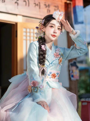 Model in a blue Hanbok adorned with 3D flowers, posing outdoors in sunlight.