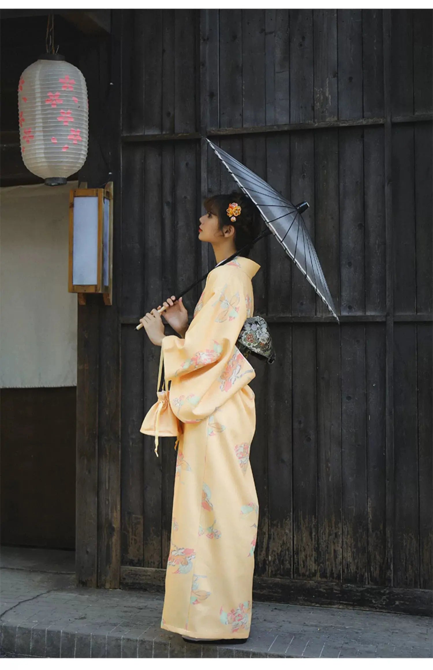 Side profile of a woman in an orange Japanese kimono-style yukata with floral designs, holding a black umbrella in a traditional setting.