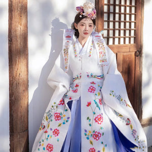 Bride in embroidered Korean hanbok with vibrant floral motifs, standing by a door.