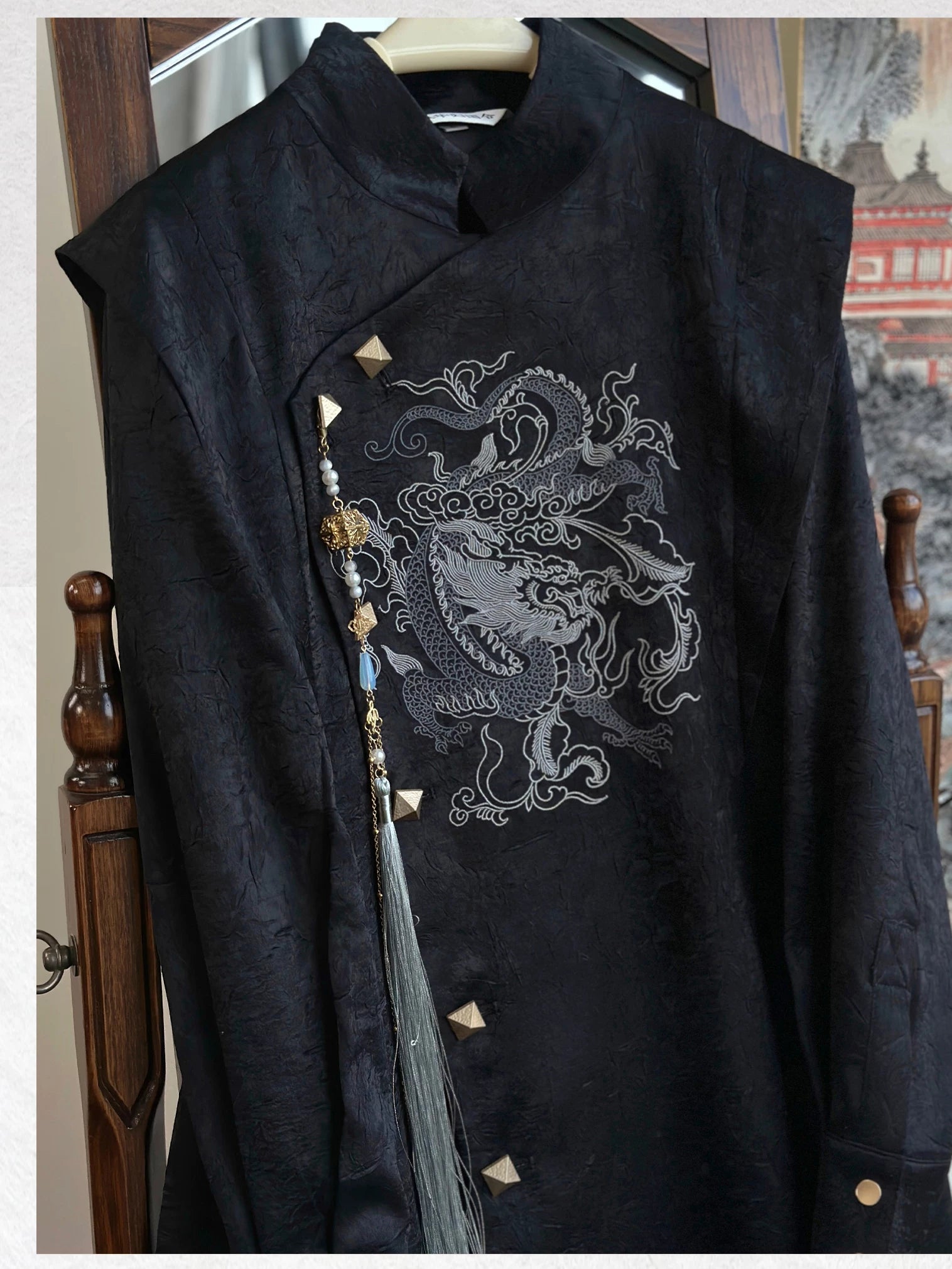 Close-up of the black dragon embroidery coat on a hanger, showing the textured fabric, asymmetric gold-tone buttons and detachable tassel.