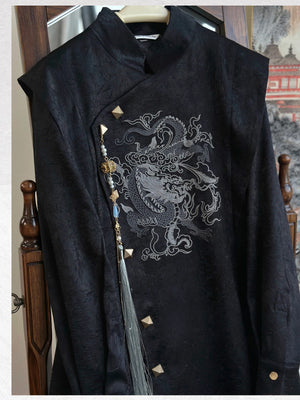 Close-up of the black dragon embroidery coat on a hanger, showing the textured fabric, asymmetric gold-tone buttons and detachable tassel.